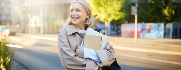 Photo candid young woman on her way to university standing on street carries notebooks and homework has