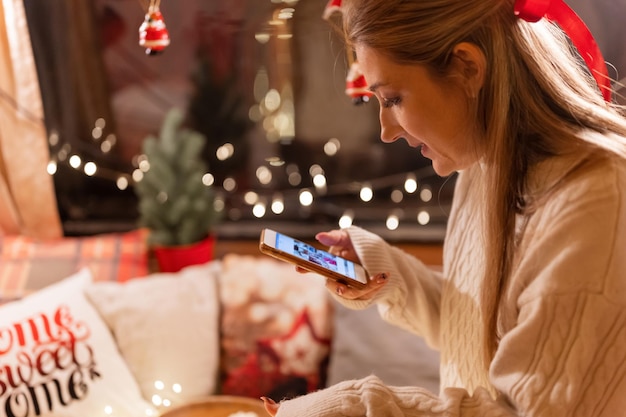 Giovane donna schietta che tiene in mano lo smartphone e guarda o visualizza le foto sul display. divertimento rilassante in campeggio in un letto accogliente durante la notte di buon natale e buon capodanno