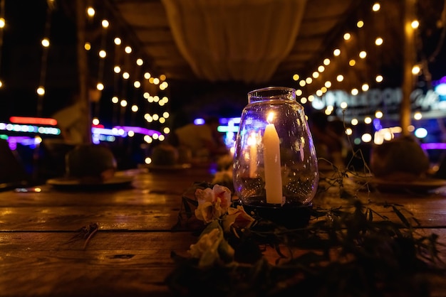 A candle in a glass jar sits on a table in front of a string of lights.