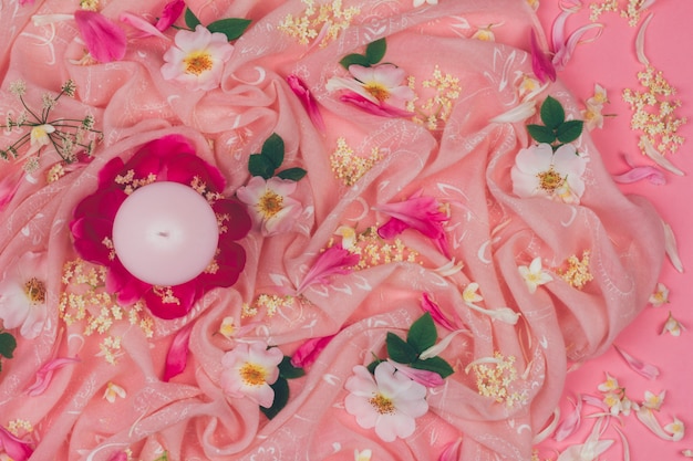 Candle and pink wild rose flowers on pink fabric background, flat lay top view