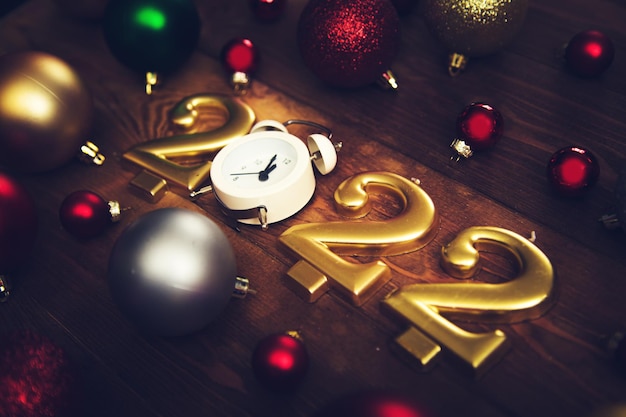 Candles in the form of numbers 2022, alarm clock showing midnight, multicolored christmas balls on a wooden background. Dark Christmas background.