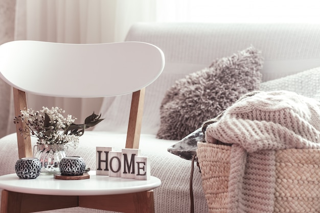 Candles, a vase with flowers with wooden letters of the home on wooden white chair. Sofa and wicker basket with pillows.