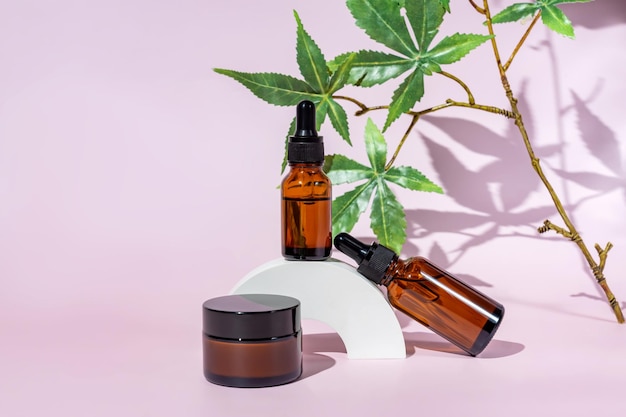 Cannabis gezichtscrème en serum of olie druppelaar concept Natuurlijke cosmetische CBD olie THC tinctuur en hennep bladeren op een roze achtergrond