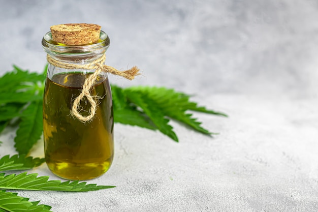 Olio di cannabis in una bottiglia trasparente e foglie di canapa su uno sfondo grigio.