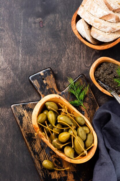 Canned capers in wooden dish, tapenade and bread on trendy dark wooden old background. Top view