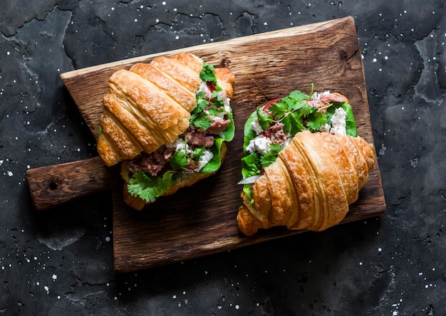 Canned tuna cream cheese tomato lettuce croissants sandwiches on rustic cutting board on a dark background top view