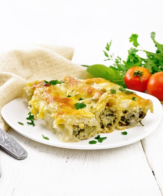 Cannelloni stuffed with cottage cheese and spinach with Bechamel sauce in a plate, napkin, tomatoes and parsley on a wooden board background