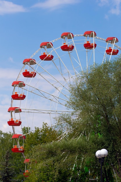 Cantilevered observation wheel with empty red cabins on blue sky and green trees in park