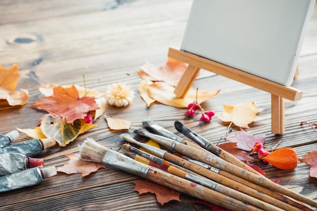 Canvas on easel paint tubes brushes for painting and autumn leaves on old wooden background