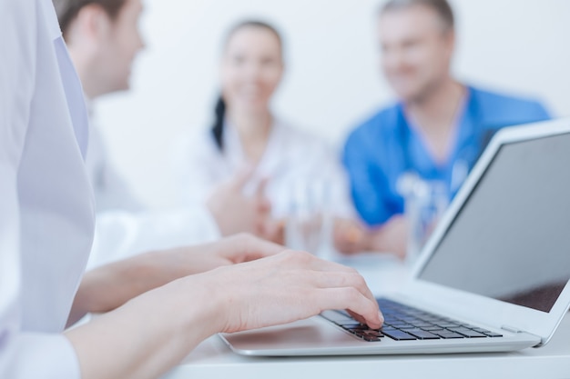 Capable professional young medic using laptop at the clinic and typing while her colleagues having discussion behind