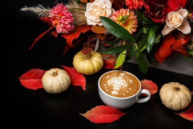 Cappuccino and beautiful flowers bouquet still life. Flower shop composition. Coffee cup, pumpkins on black wooden table. Florist art and floral design concept