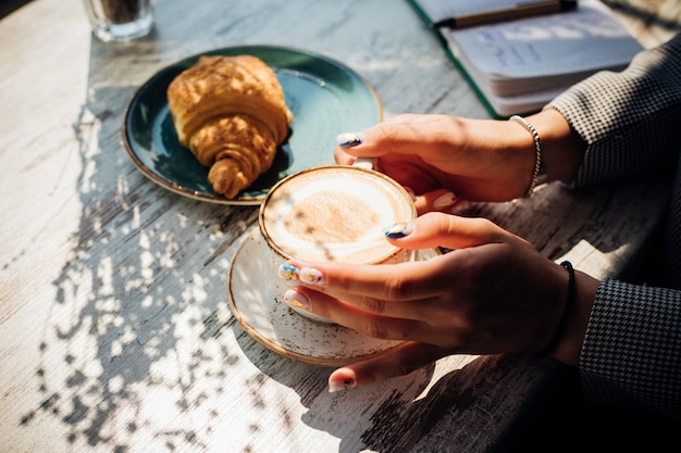 Cappuccino en croissant op tafel in het café. Het ochtendzonnetje valt op tafel, er verschijnen prachtige schaduwen. Heerlijk ontbijt. De handen van vrouwen houden een kopje koffie vast.