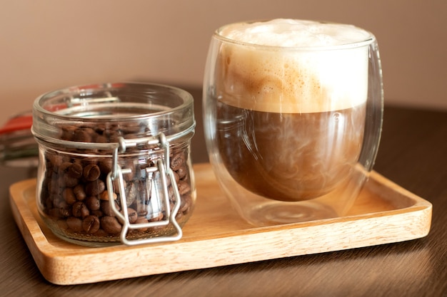 Капучино в двойной стеклянной чашке и банке с кофейными зернами на деревянной тарелке.