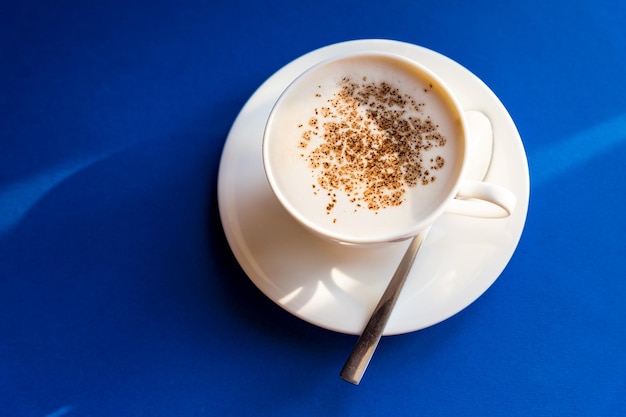 Cappuccino op blauwe achtergrond witte kop met lepel koffiebonen 's ochtends koffie in Italië Traditioneel Italiaans ontbijt Kleine witte keramische kopjes met koffie Copz ruimte