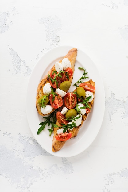 Caprese bruschetta toast met kerstomaatjes, mozzarella, olijven en basilicum op wit bord oud licht oppervlak. Bovenaanzicht.