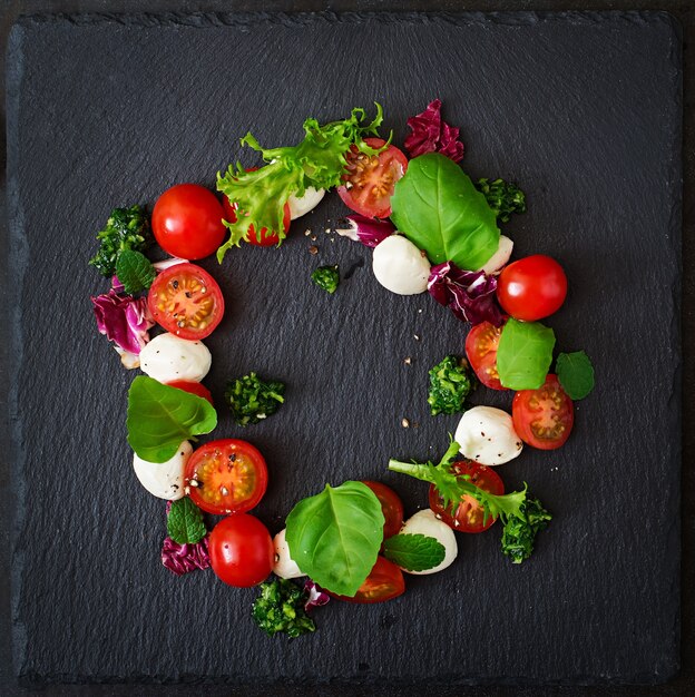 Photo caprese salad tomato and mozzarella with basil and herbs oon black background. top view