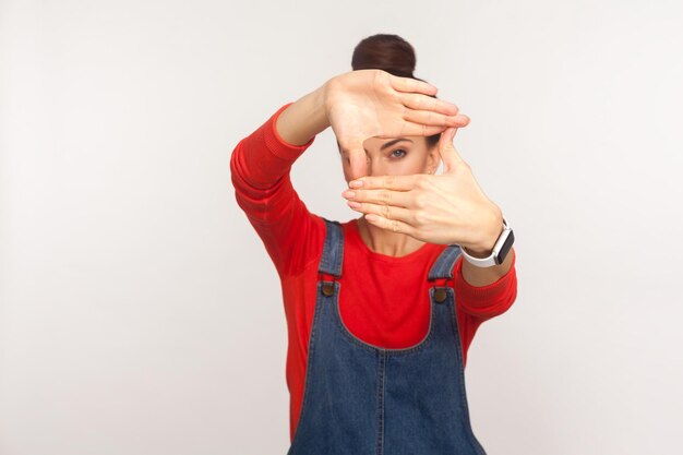 Foto cattura momenti. ritratto di ragazza in tuta di jeans che guarda attraverso la forma della cornice per foto con le dita, osserva da lontano, mette a fuoco e ingrandisce la fotocamera. tiro in studio indoor isolato su sfondo bianco