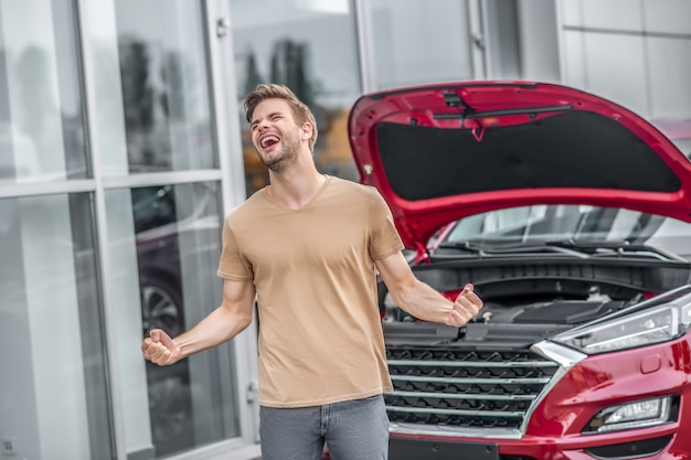 車の故障。絶望的な男性ドライバーが叫び、ボンネットを開けた壊れた車の前で拳を握り締める