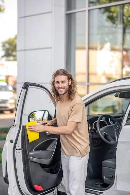 Car care. smiling long-haired friendly guy in casual clothes\
wiping front door of car with yellow napkin outdoors on sunny\
day
