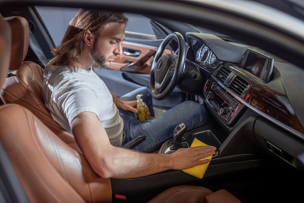 Car care. Young bearded serious enthusiastic man carefully wiping surfaces while sitting in car in drivers seat