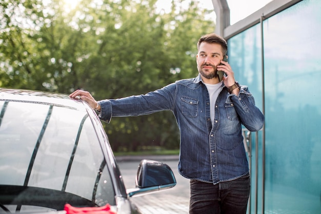 セルフサービスの洗車場で車の近くに立ち、スマートフォンで話している若い白人男性のポートレート