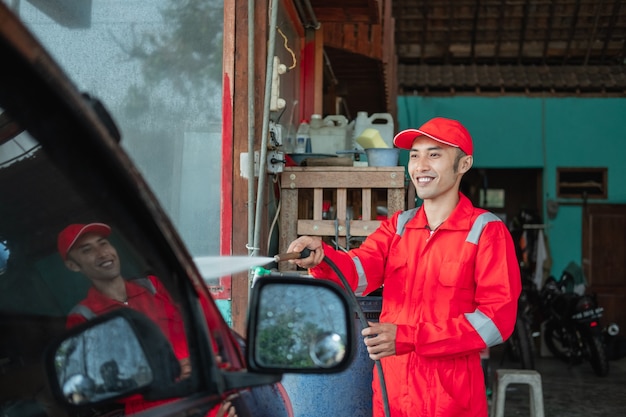 A car cleaner wearing a red uniform and a standing hat sprays water using a hose to the car in the car wash