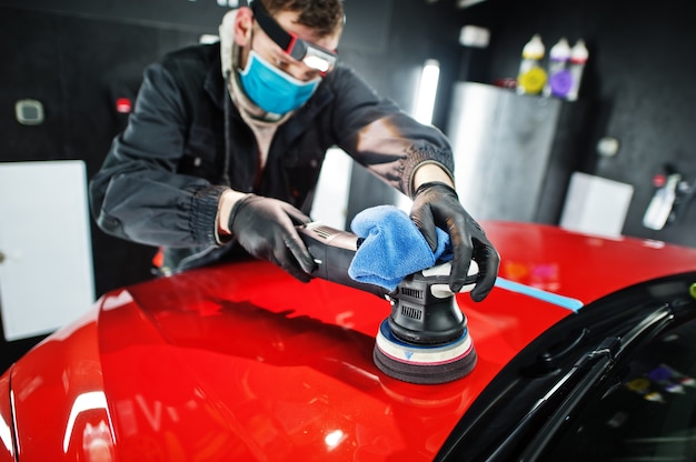 Photo car detailing concept. man in face mask with orbital polisher in repair shop polishing orange suv car.