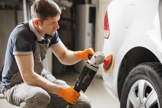 Car detailing male worker holds a polisher in the hand and\
polishes white car in auto repair shop