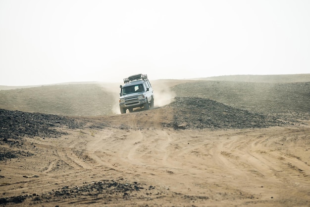 Автомобиль проезжает через пыльные песчаные барханы в пустыне на фоне белого неба, сафари-поездка на джип-дюне б ...