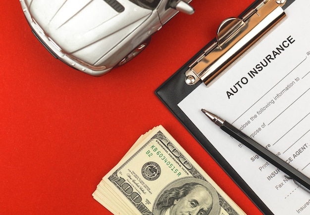 自動車保険フォーム。エージェントのオフィスビジネスデスクトップ上の合意とポリシー、お金と車のおもちゃのクリップボード。赤い背景と上面写真