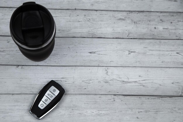 A car key with automatic buttons and a thermocup are black with metal inserts on a white wood