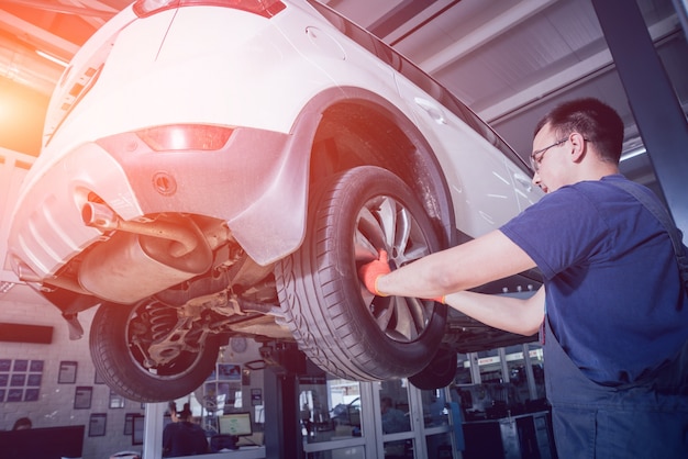 Meccanico di automobile che ispeziona dettaglio della ruota e della sospensione dell'automobile sollevata al distributore di benzina di riparazione