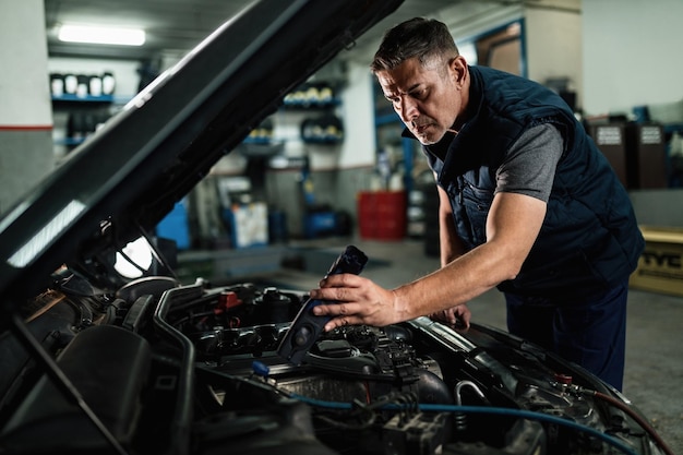 Car mechanic using lamp while examining engine under the hood at auto repair shop