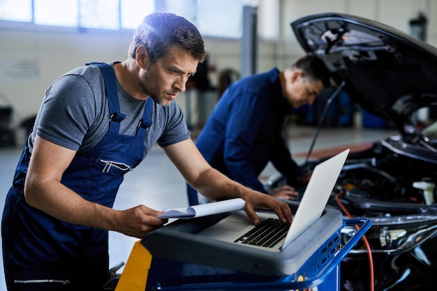 Car mechanic using laptop while analyzing car diagnostic in\
auto repair shop his coworker is in the background