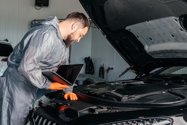 Meccanico di automobile che lavora con un computer portatile nel servizio di riparazione automatica che controlla il motore dell'automobile