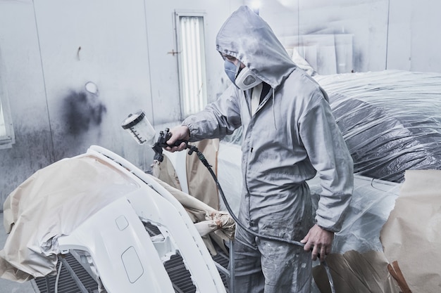 Car painting and automobile repair service. Auto mechanic in white overalls paints car with airbrush pulverizer in paint chamber