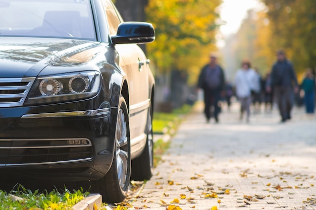 Car parked on a city street side on bright autumn day with\
blurred people walking in pedestrian zone