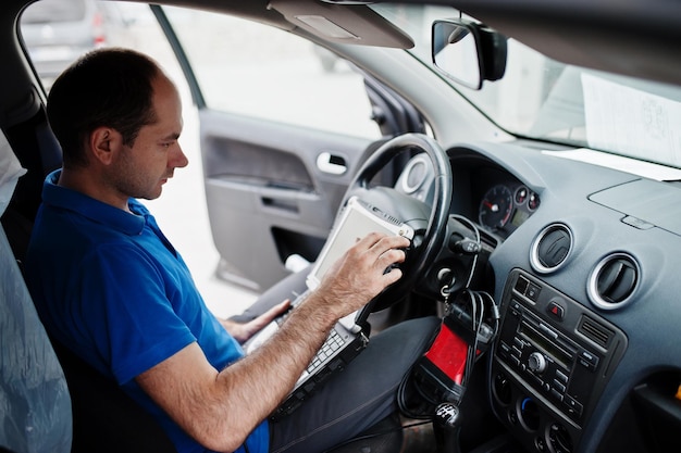 Car repair and maintenance theme. Electric mechanic in uniform working in auto service, making car diagnostics using obd device with laptop.