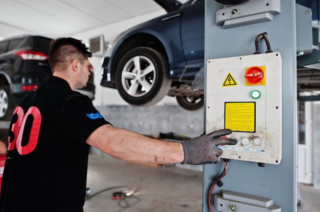 Car repair and maintenance theme. Mechanic in uniform working in auto service, press button to lifting auto.