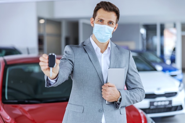 Venditore di auto con maschera facciale in piedi nel salone dell'auto e mostrando le chiavi su una nuova auto di marca che sono pronte per essere vendute