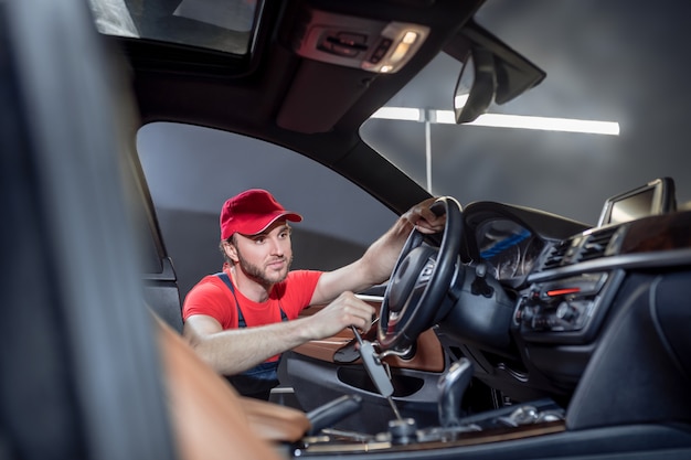 Car service. Experienced knowledgeable young man in overalls doing car service holding steering wheel in cab