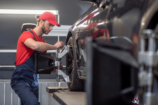 Car service. Profile of a young bearded involved man in blue overalls repairing car wheel in car workshop