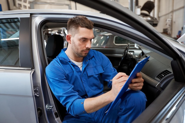 car service, repair, maintenance and people concept - auto mechanic man or smith with clipboard writing at workshop