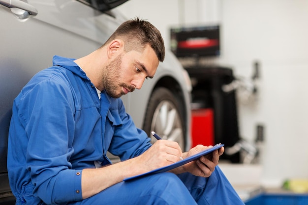 car service, repair, maintenance and people concept - auto mechanic man or smith with clipboard writing at workshop