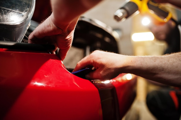 Car service worker put anti gravel film on a red car body at the detailing vehicle workshop. Car protection with special films.