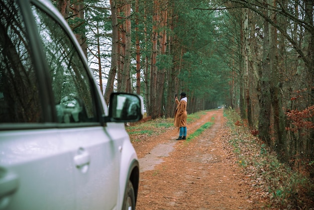 Женщина-концепт автомобильного путешествия вышла из белого внедорожника на лесной тропе, чтобы сфотографироваться на телефон