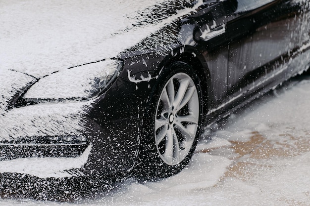 Car wash concept close up shot of dirty black car covered with\
white foam at cleaning station washing process perfect service