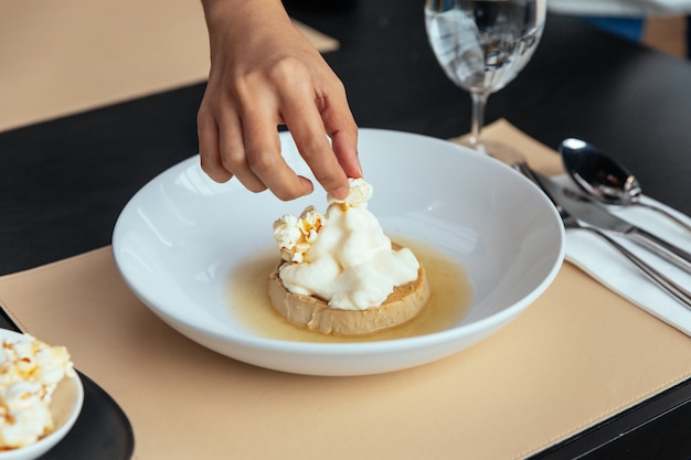 Caramel Custard met slagroom die met de hand een popcorn maakt. Geserveerd in witte plaat op lederen mat.