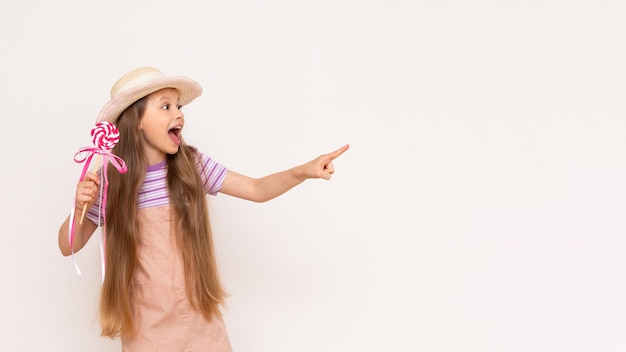 A caramel lollipop in the hands of a beautiful little girl A baby in a summer sundress and a summer hat points to a white isolated background with her index finger