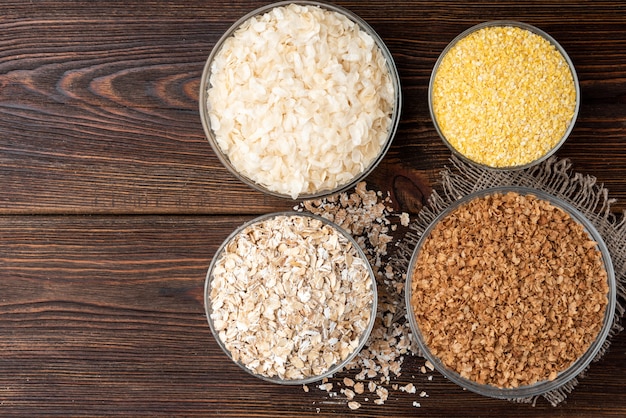 Carbohydrates. Various grain flakes in bowls on dark wooden table.